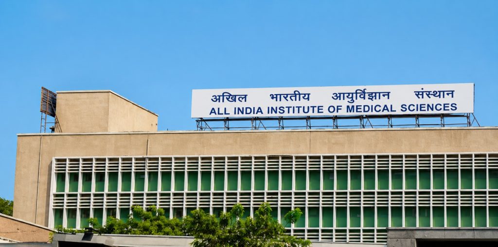 Construction of All India Institute of Medical Sciences (AIIMS) with the institute name on rooftop signage against clear blue sky.